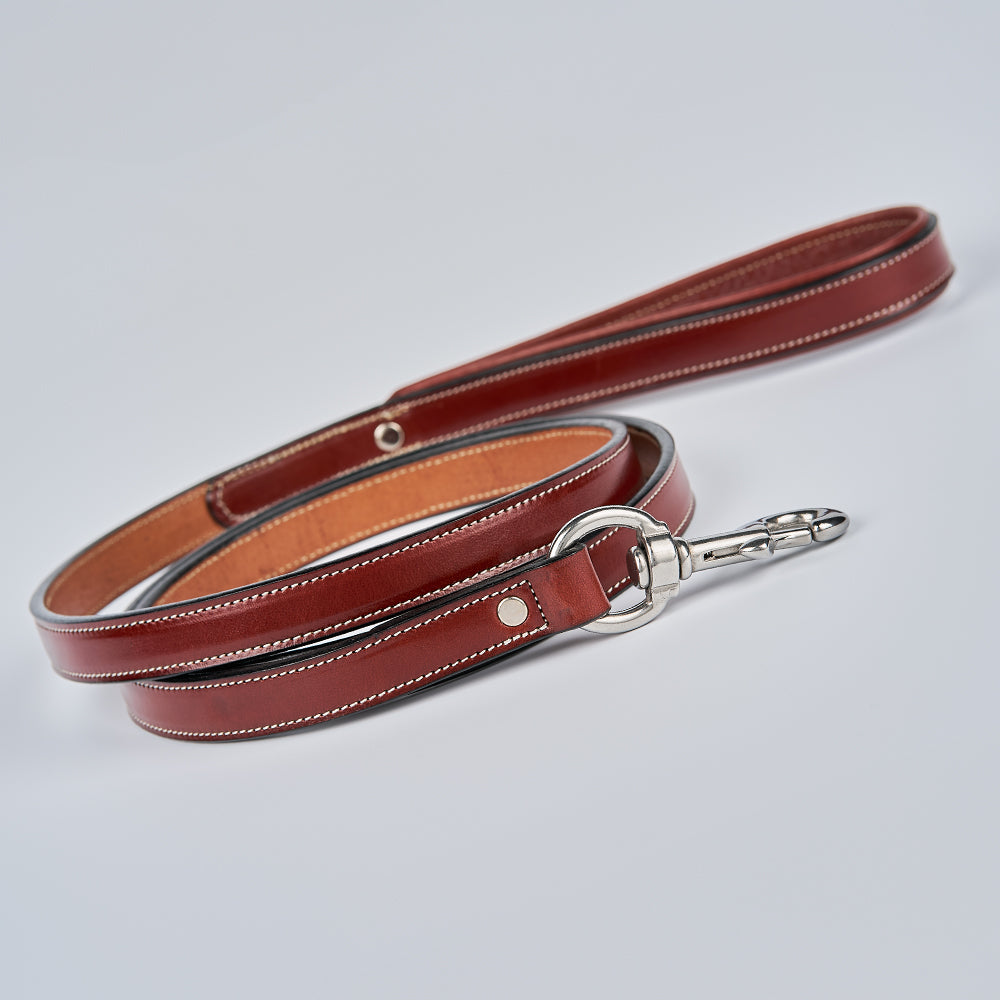 The Rover Standard Leash by Hanks Belts features brown English bridle leather, white stitching, a padded handle, and a silver clasp. It’s neatly coiled on a light gray background.