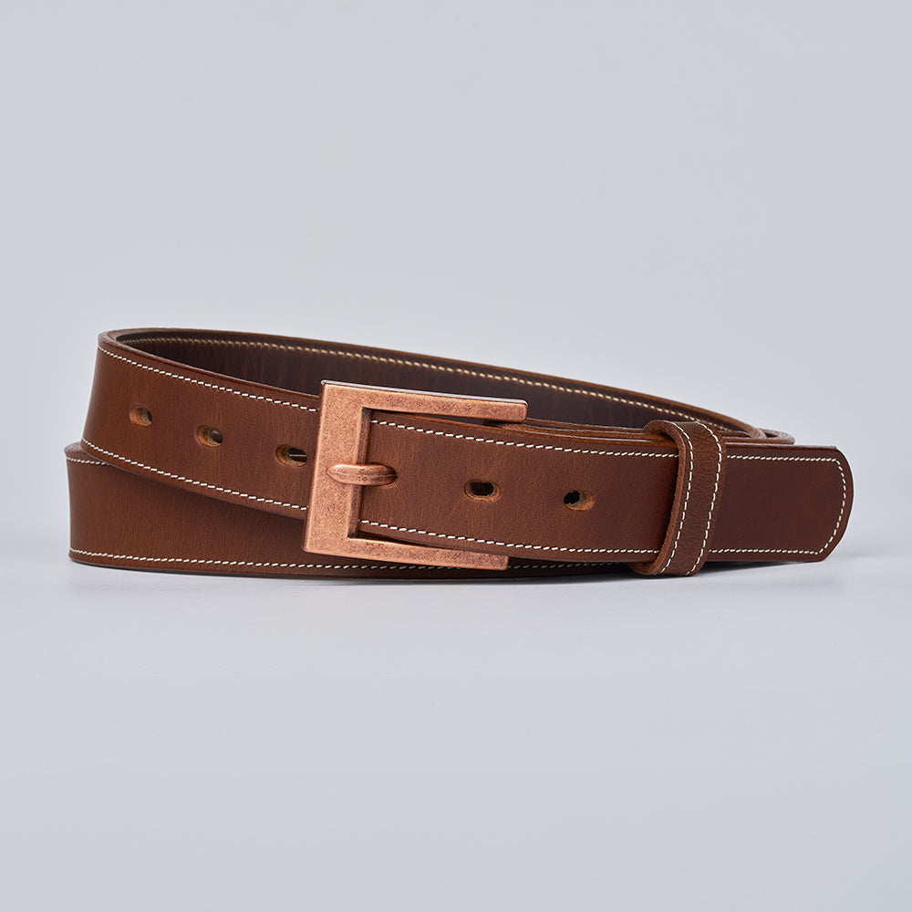 The Heritage Bison by Hanks Belts: a rugged brown bison leather belt with white stitching and a rectangular copper-colored buckle, coiled neatly on a light gray background—showcasing timeless American craftsmanship.