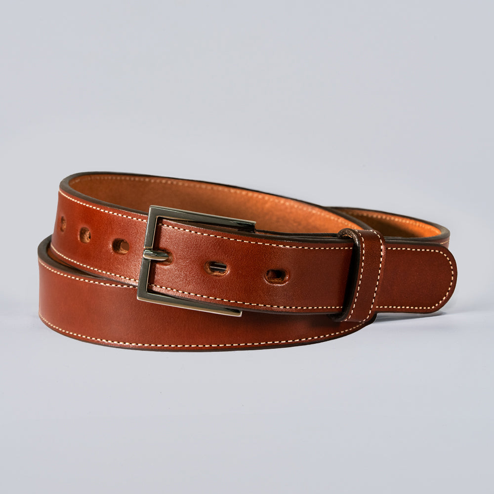 The Ledger by Hanks Belts is an oak leather belt with a silver rectangular buckle, white edge stitching, and multiple adjustment holes, coiled neatly on a light gray background.