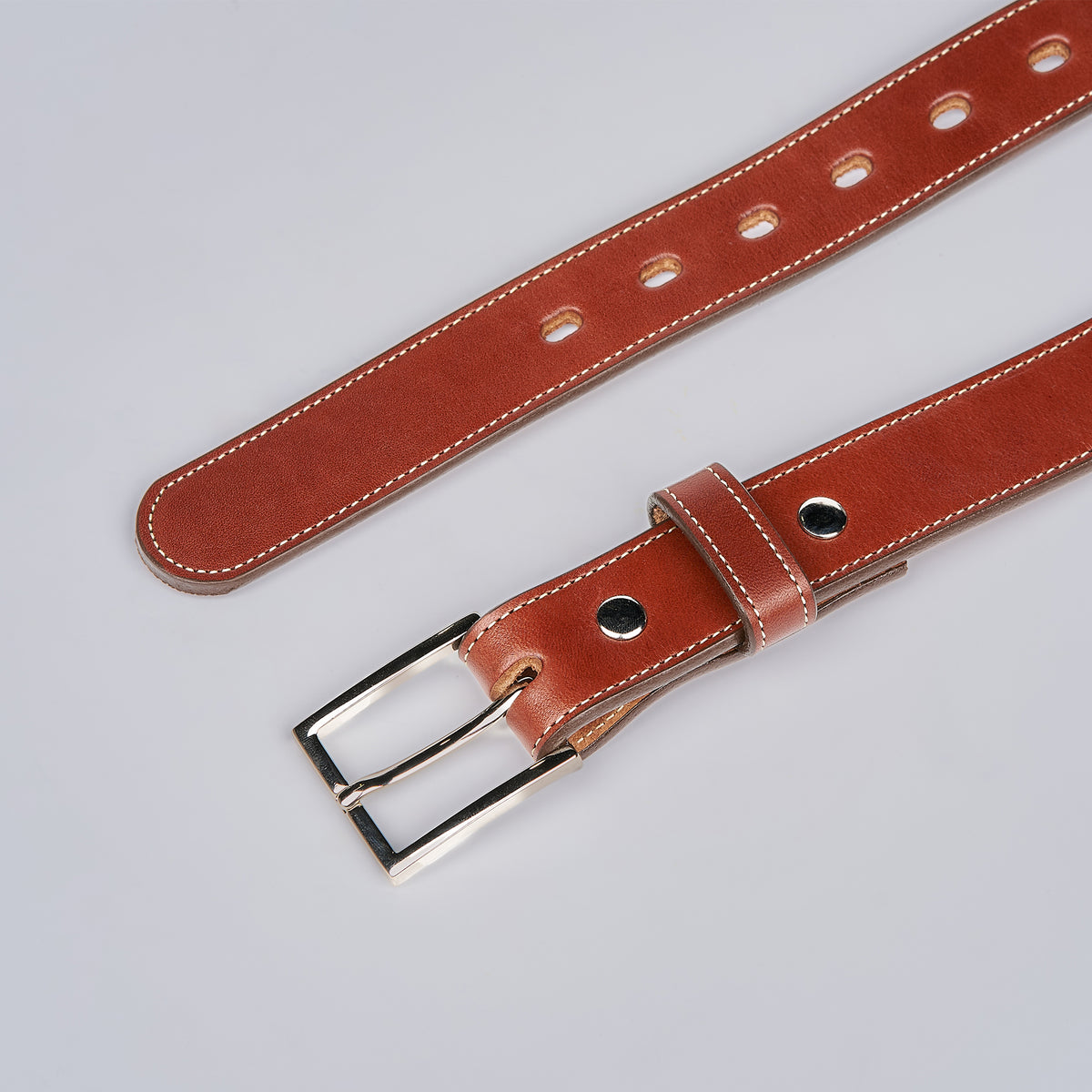 The Ledger by Hanks Belts, a brown English bridle leather belt with white stitching, silver buckle, and multiple holes, is displayed on a light grey background.