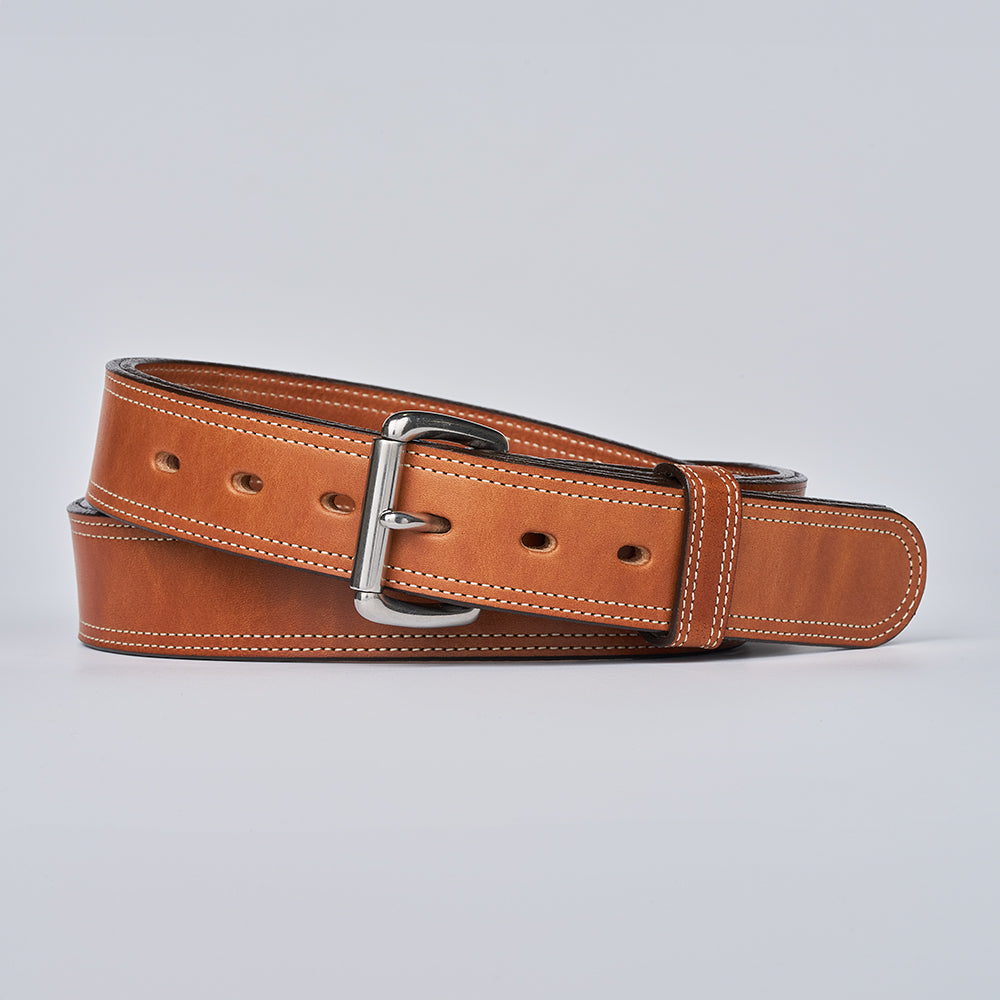 The Premier Double Layer by Hanks Belts, a tan vegetable tanned leather belt with white stitching and a silver buckle, is coiled neatly against a plain light background.