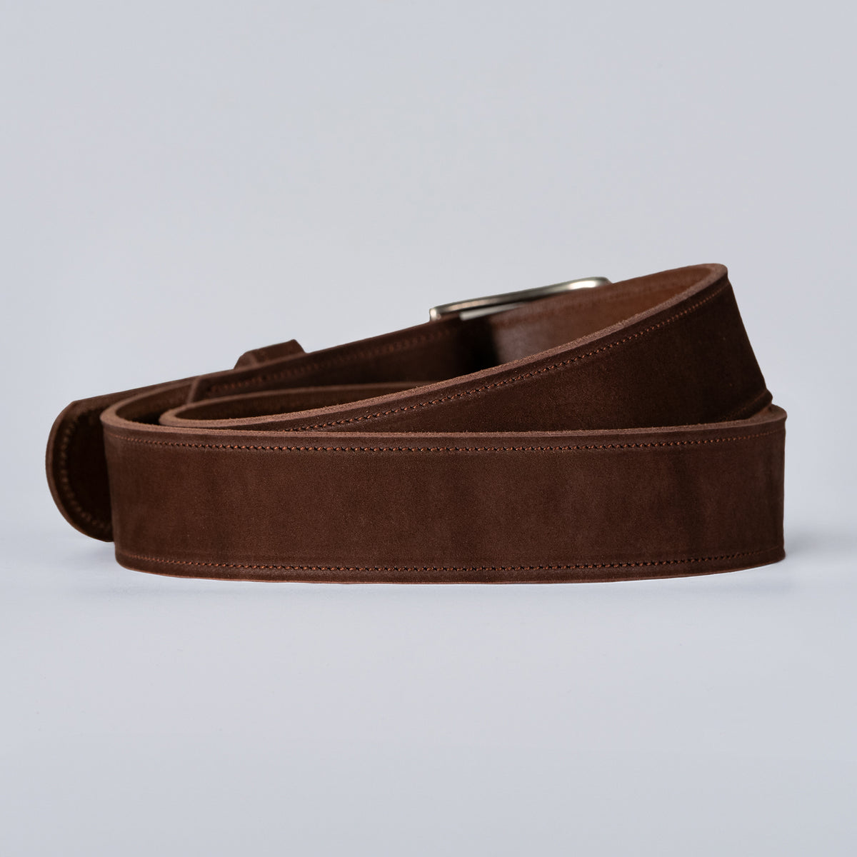 The Suede - 1.5" by Hanks Belts: a premium nubuck leather belt in brown, with a silver buckle, coiled and displayed on a plain light gray background.