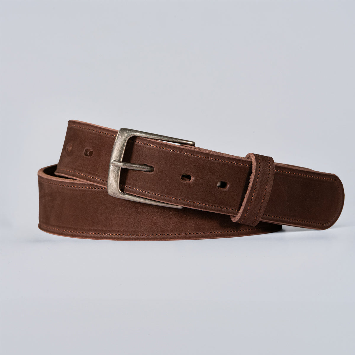 The Suede - 1.5" by Hanks Belts, a brown USA-made suede belt with a silver buckle, is coiled neatly on a light gray background.