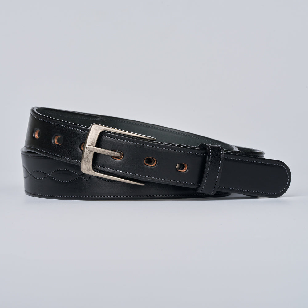 The Teton Tapered by Hanks Belts is a black handcrafted leather belt with decorative stitching, multiple eyelets, and a silver buckle, rolled up on a plain light gray background.