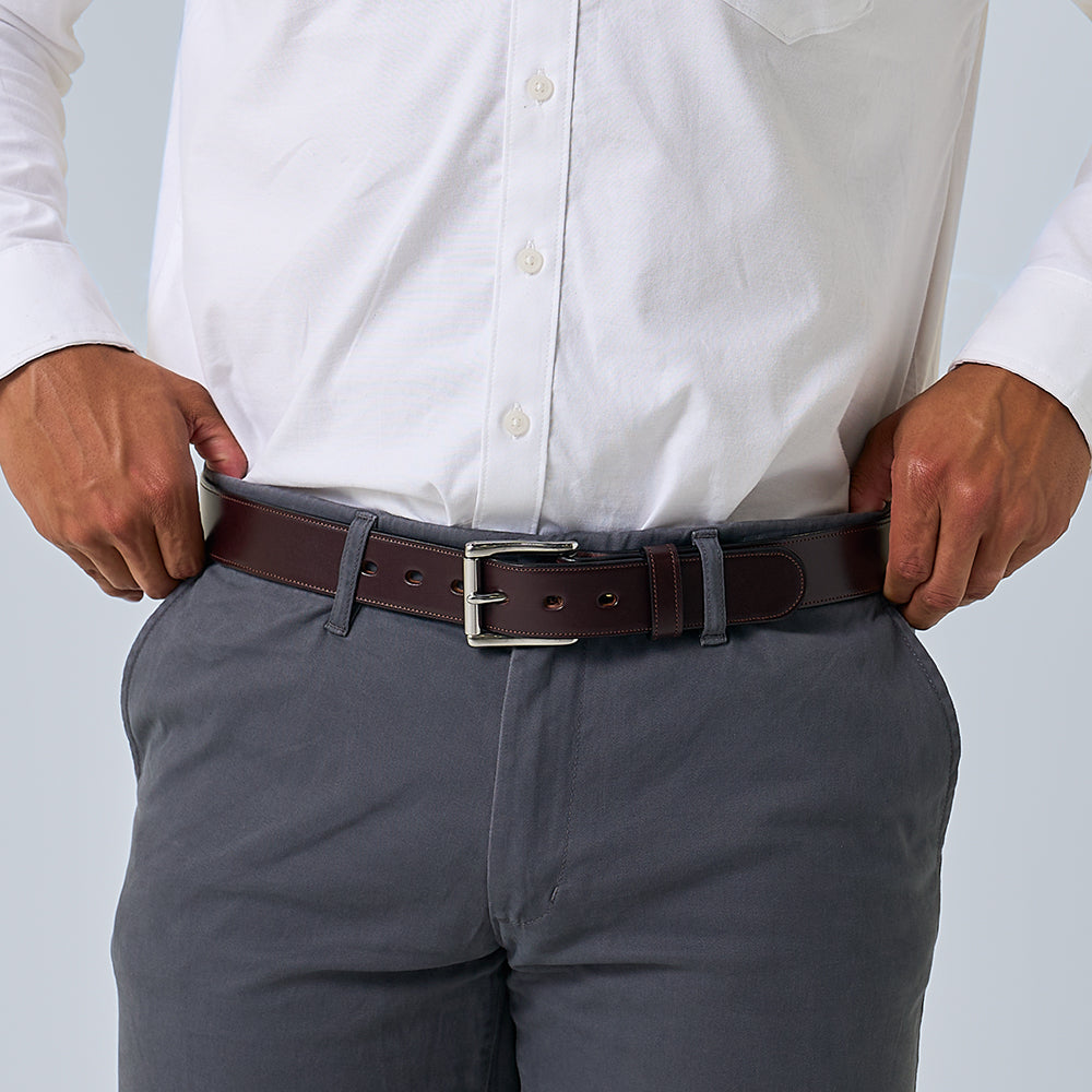 Wearing a white button-up shirt tucked into gray pants, a person adjusts The Canyon by Hanks Belts—a handcrafted dark brown vegetable-tanned leather belt with a silver buckle. Only the torso and hands are visible.