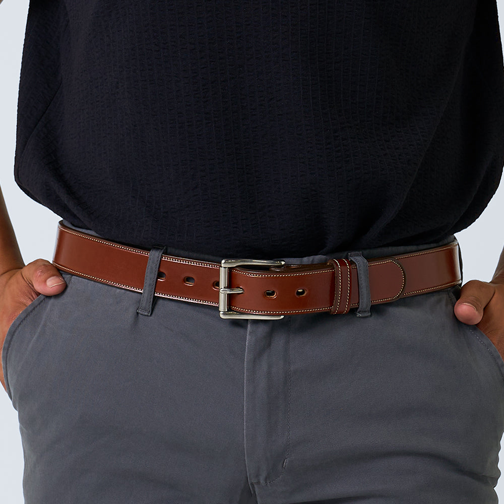 A person stands with hands in their pockets, wearing a dark textured shirt, gray pants, and The Canyon handcrafted English bridle leather belt with a silver buckle by Hanks Belts.