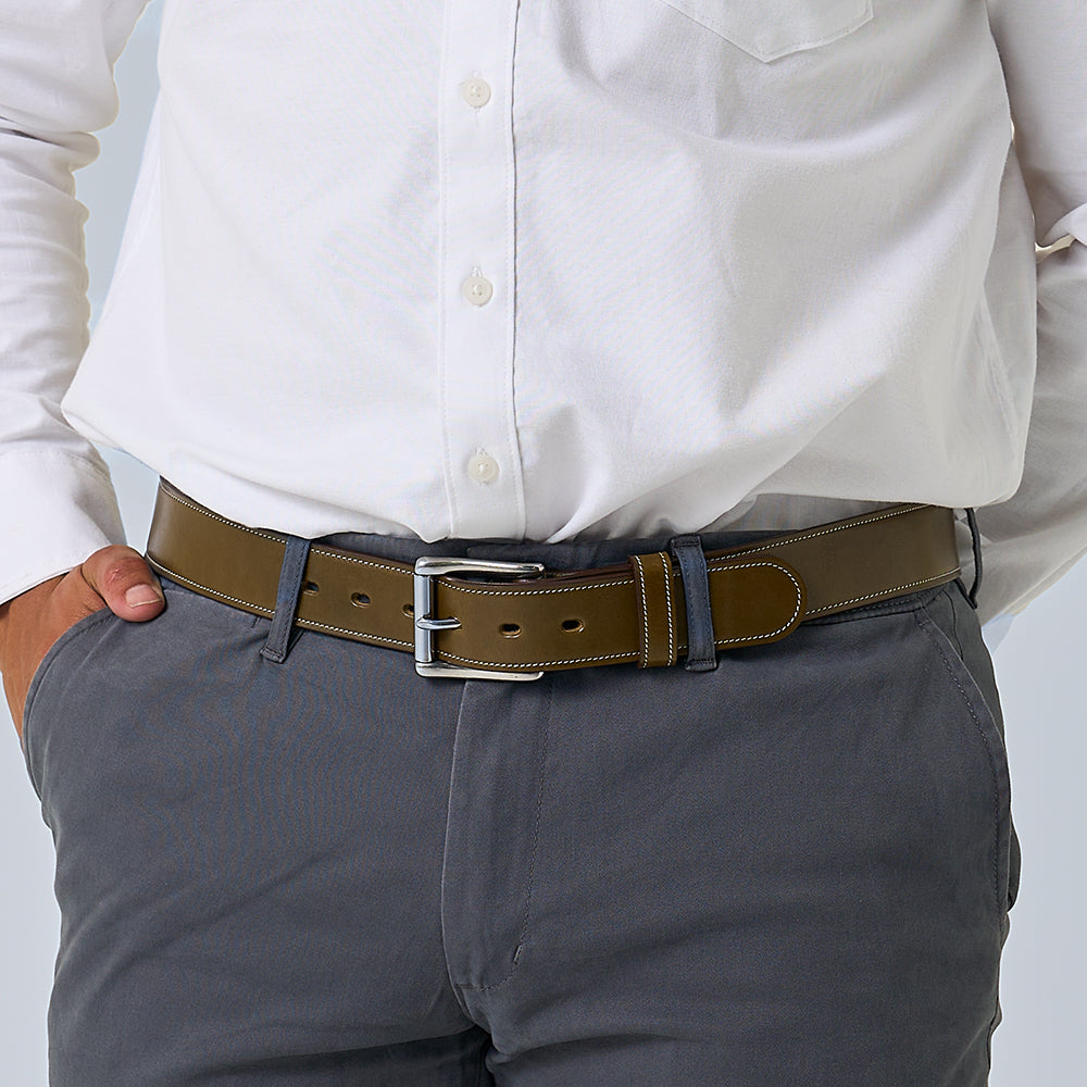 A person wearing a white button-up shirt, gray pants, and The Canyon handcrafted vegetable-tanned leather belt by Hanks Belts, standing with one hand in their pocket.