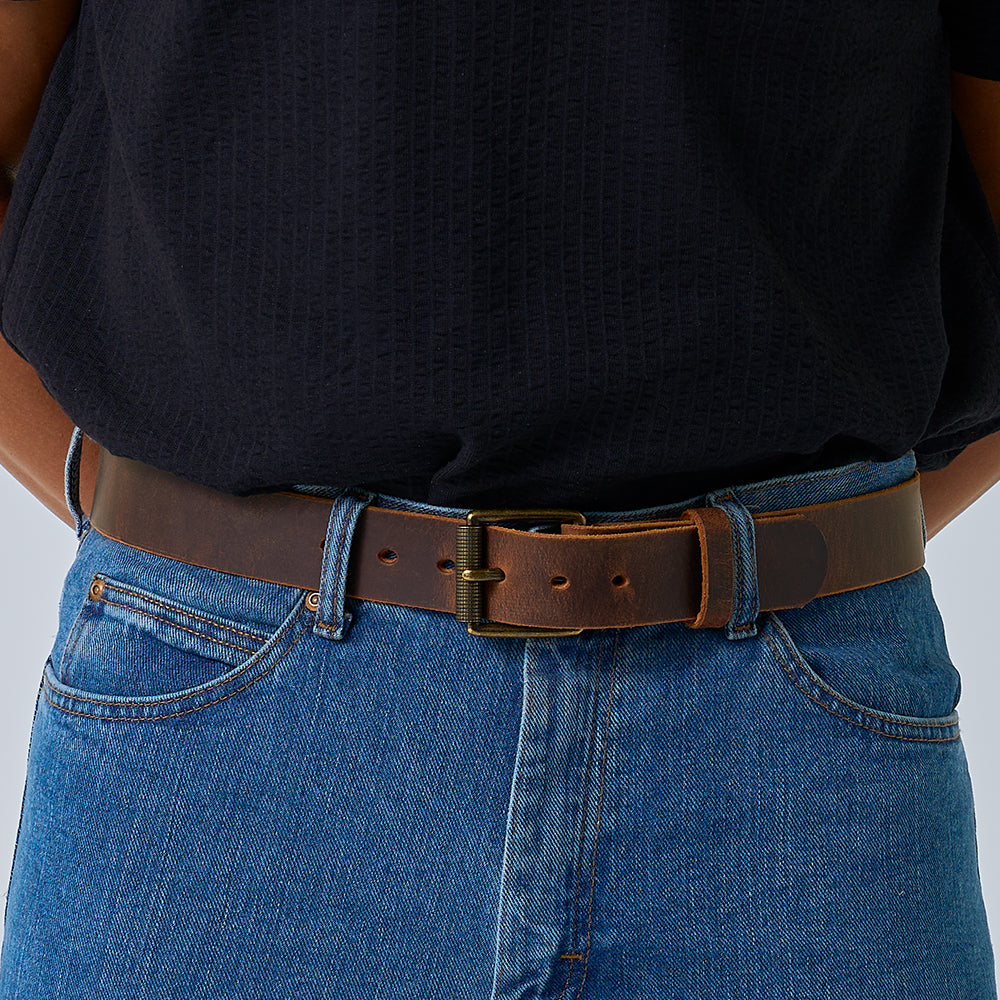 A person in a textured black shirt and blue jeans wears Hanks Belts’ The Crazy Horse, a USA-made belt with dark brown Water Buffalo leather and brass buckle, arms behind their back.
