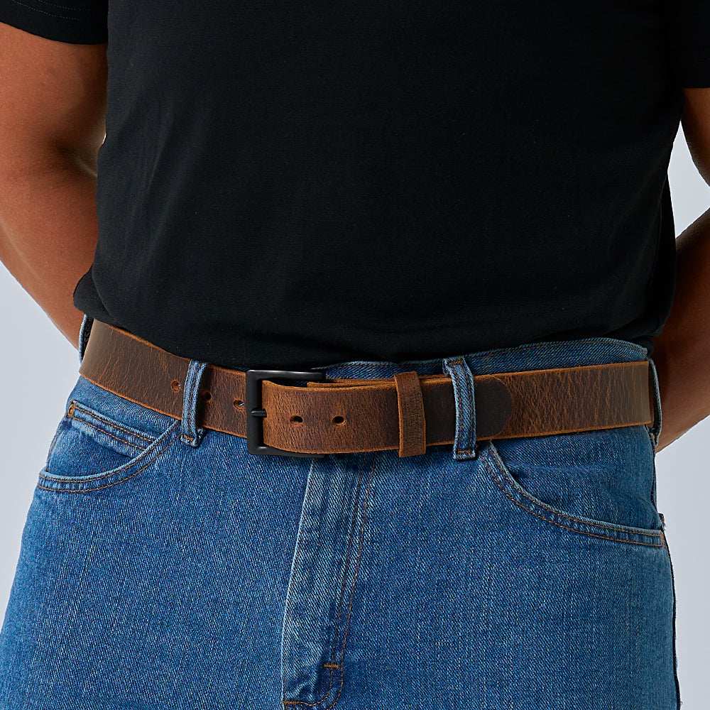 A person wears a black t-shirt tucked into blue jeans, secured with The Crazy Horse belt by Hanks Belts, featuring a retro leather design and black buckle. Their hands are behind their back.