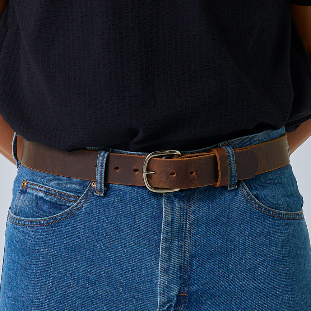 A person wearing blue jeans and a black textured shirt showcases The Crazy Horse leather belt by Hanks Belts, fastened with a metal buckle, shown from the waist down.