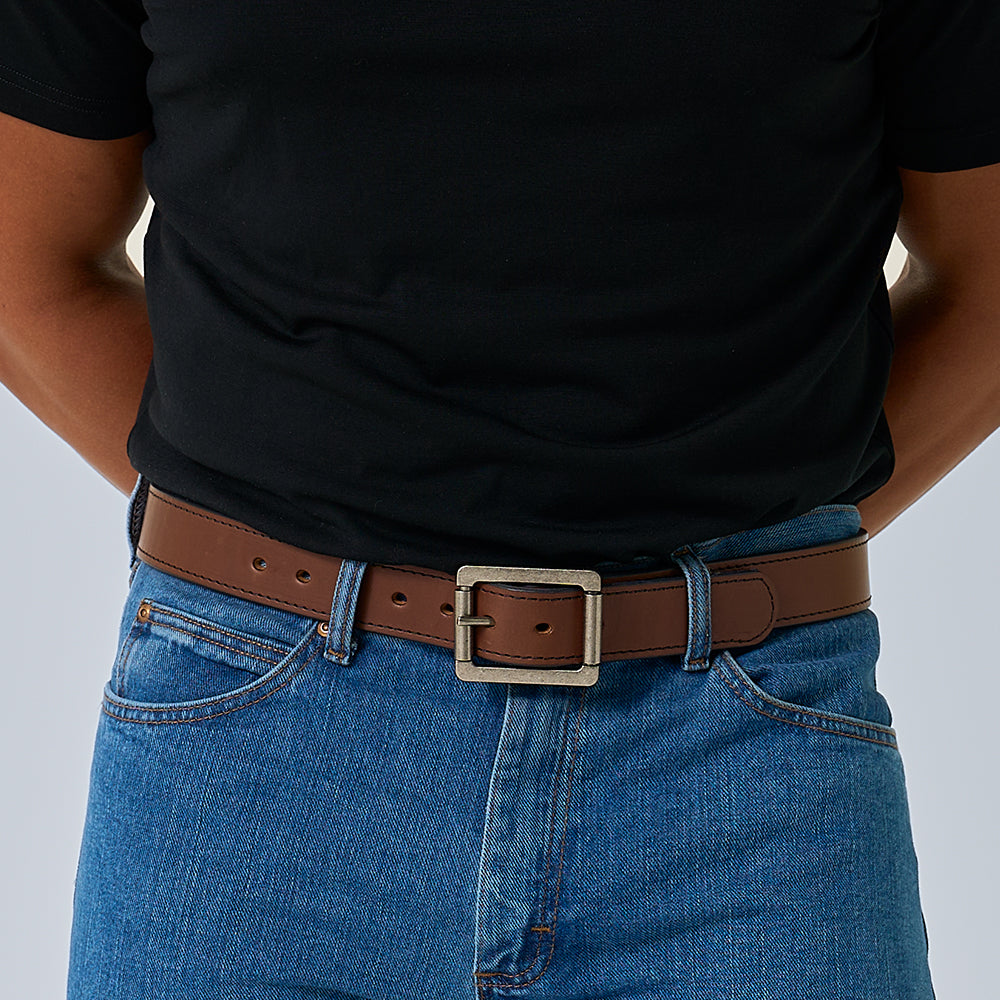 A person in a black t-shirt, blue jeans, and The Double Roller by Hanks Belts—a brown full-grain leather belt with a large silver buckle—stands with hands behind their back.