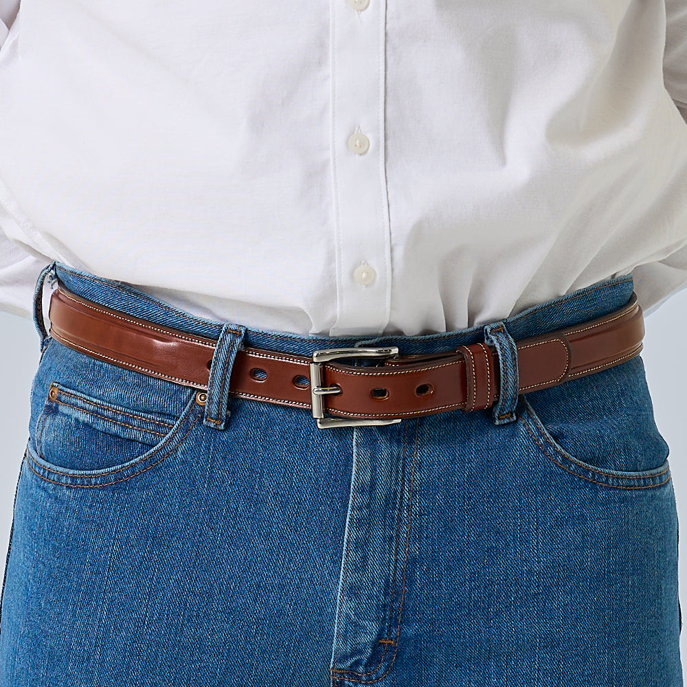 A person wearing blue jeans and a white shirt tucked in showcases the Hanks Belts Esquire Raised, an English Bridle leather belt with a silver buckle. The image highlights the waist, focusing on the belt’s premium quality.
