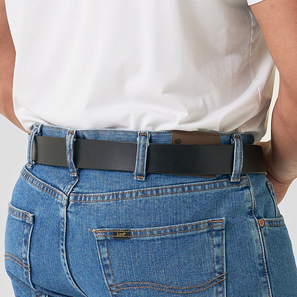 A person wears a white shirt tucked into blue jeans, showcasing Hanks Belts' "The Everyday" full-grain leather belt. Only their lower back and waist are visible, with a small "Lee" tag on the jeans pocket.