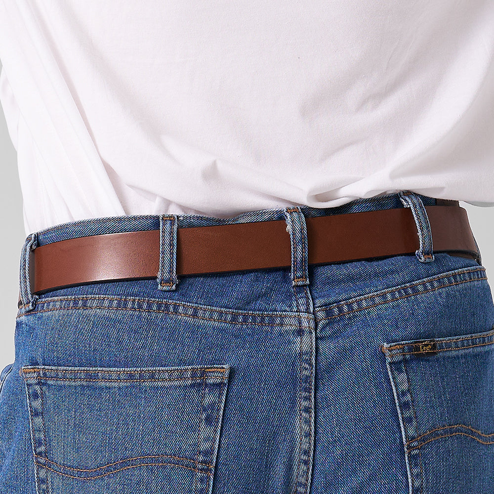 A close-up shows a person wearing Hanks Belts' The Everyday full-grain leather belt with blue jeans and a tucked-in white shirt, highlighting the back waist and pockets.