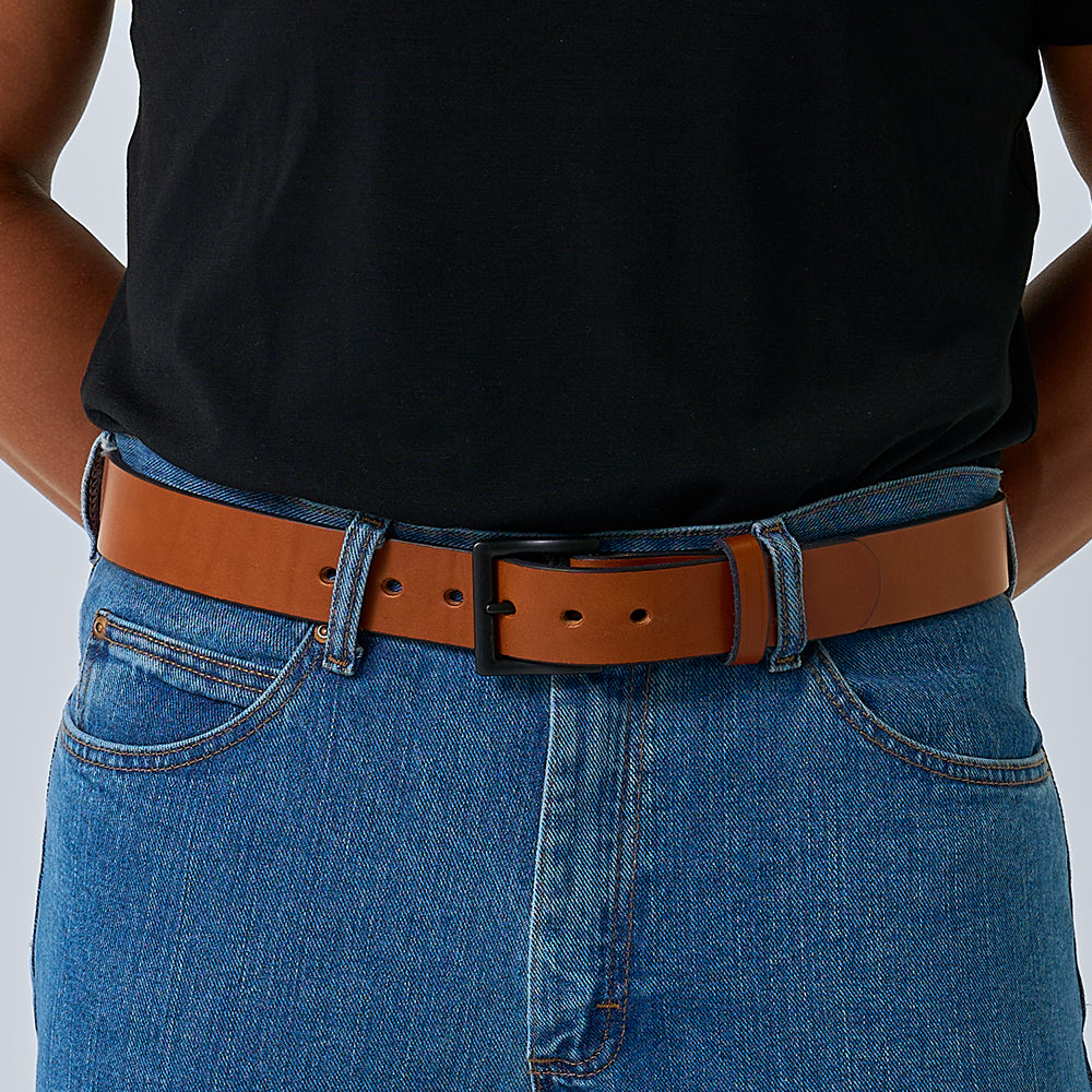 A person wears a black t-shirt tucked into blue jeans, secured with Hanks Belts' The Everyday full-grain leather belt featuring a black rectangular buckle. Their hands are positioned behind their back.