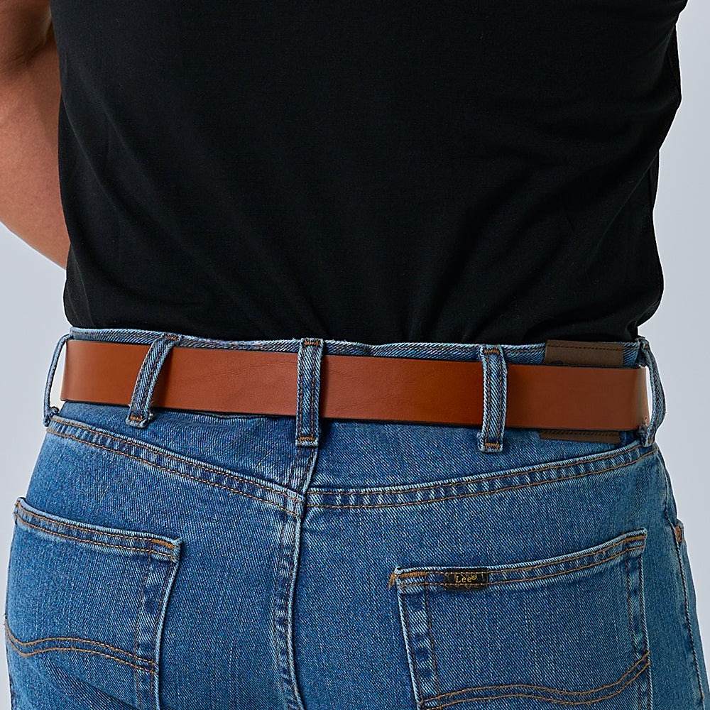A person seen from the back wears blue denim jeans with visible stitching and pockets, paired with Hanks Belts' "The Everyday" full grain leather belt and a black shirt.
