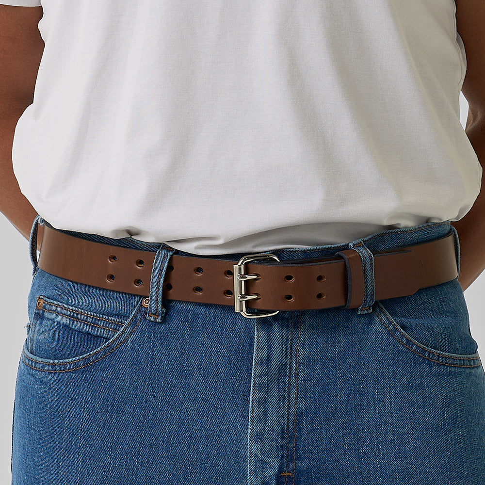 A person wearing a white t-shirt tucked into blue jeans, accessorized with Hanks Belts' The Double Prong Everyday—a brown full-grain leather belt featuring a retro double prong silver buckle.
