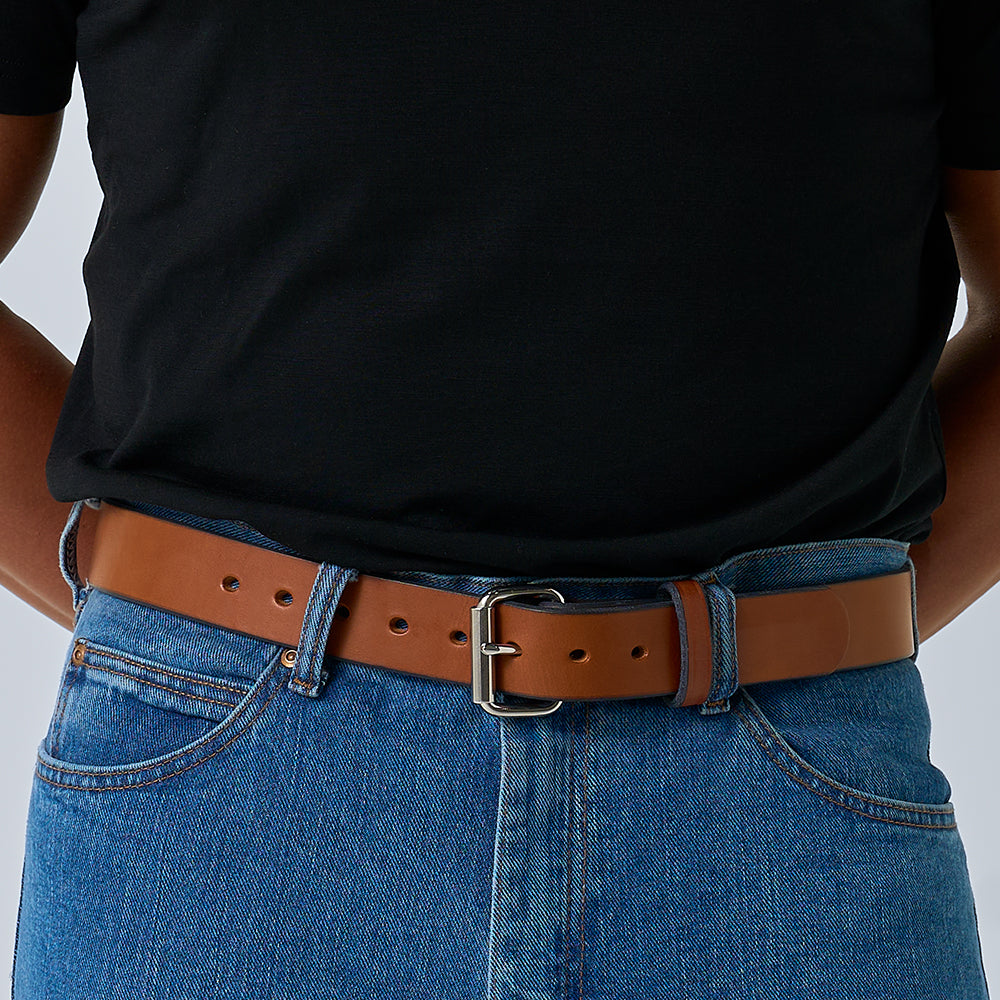 A person in a black t-shirt and blue jeans wears The Gunner by Hanks Belts, a durable work belt with a brown leather strap and silver buckle.