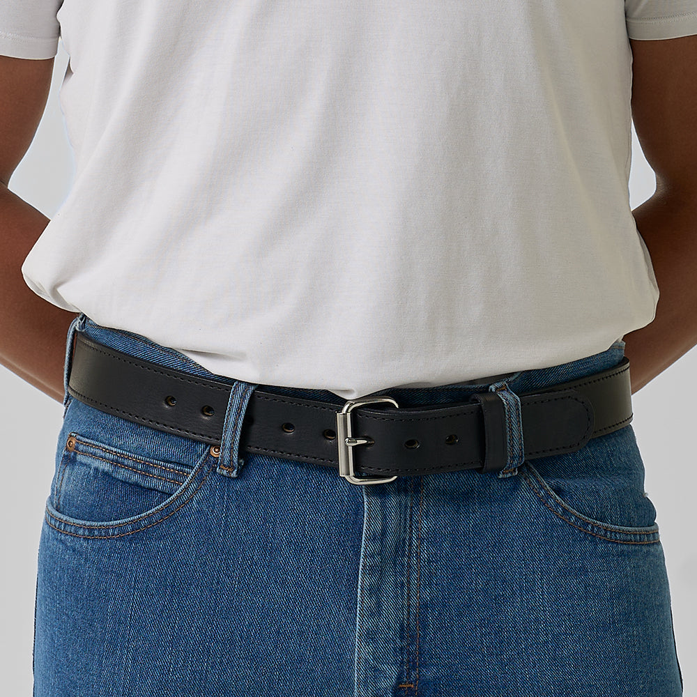 A person wears a plain white t-shirt tucked into blue jeans, secured with The Gunner Stitched belt by Hanks Belts—a black full-grain leather CCW belt with a silver buckle. Their hands are behind their back.