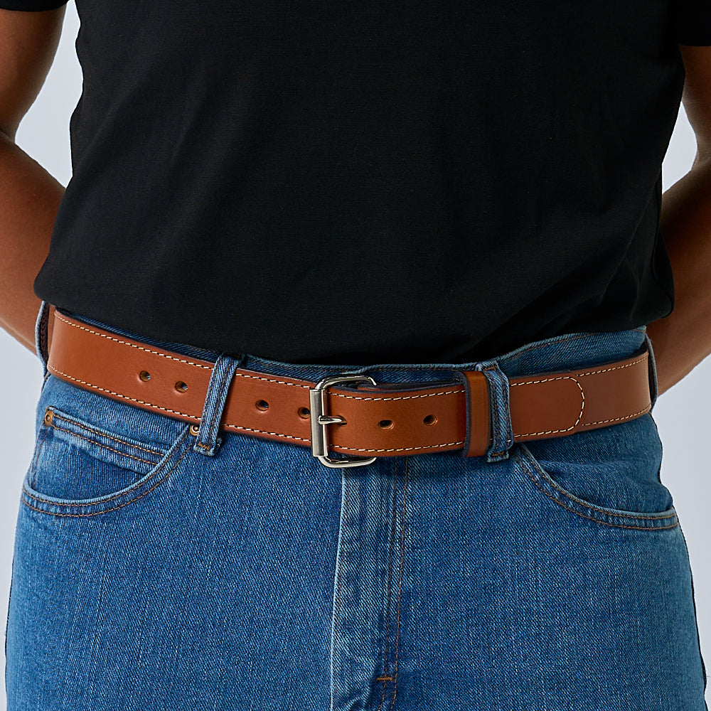 A person wears a black T-shirt tucked into blue jeans with Hanks Belts’ The Gunner Stitched, a brown full-grain leather gun belt with a silver buckle. Their hands are resting behind their back.