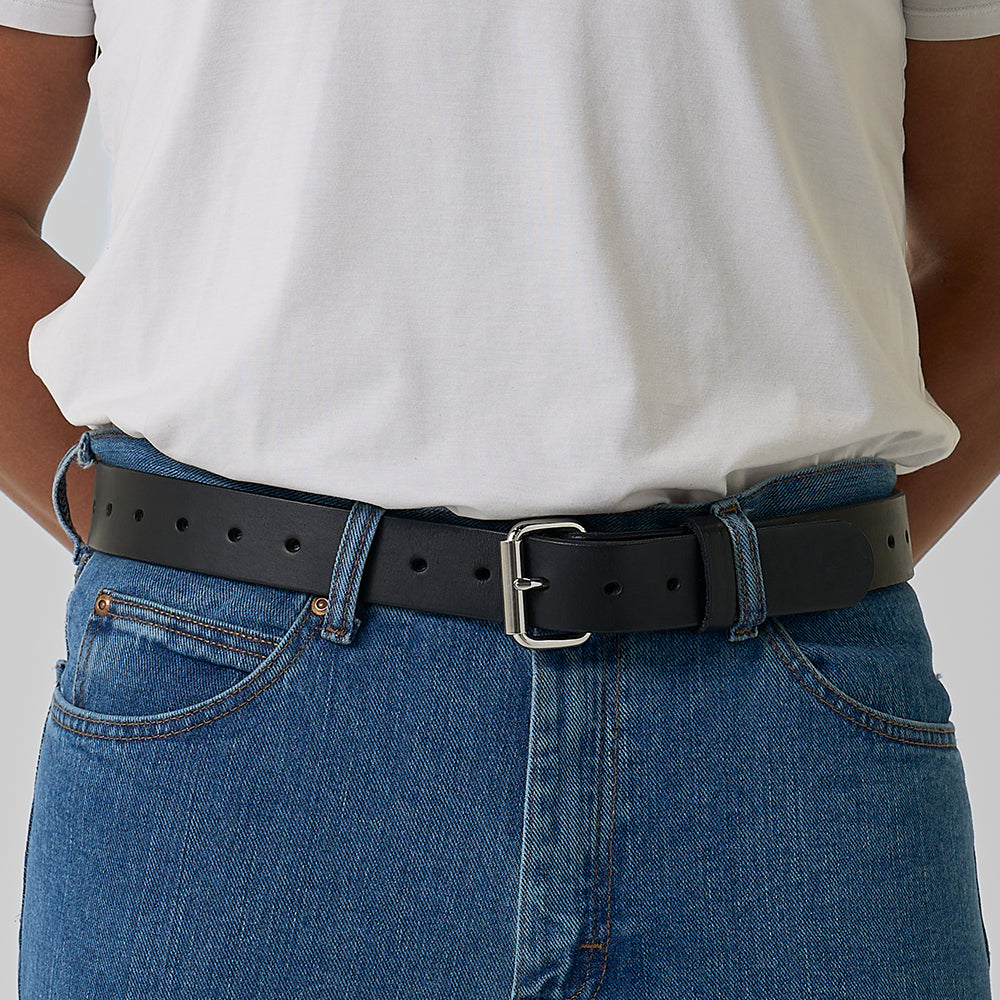 A person wearing a white t-shirt, blue jeans, and The Gunner Utility belt by Hanks Belts with a silver buckle stands with hands in their pockets.