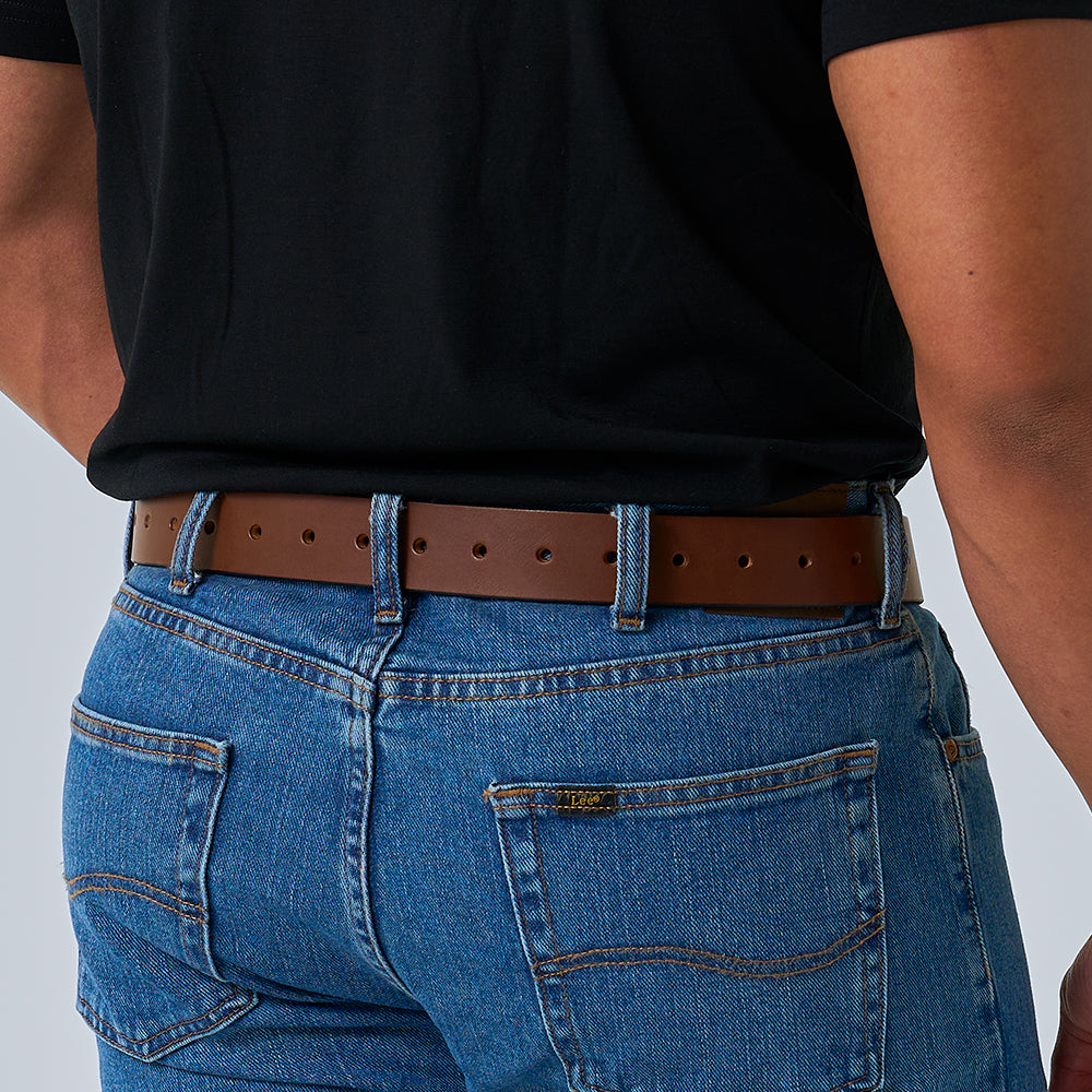 A person wearing blue jeans with double-stitched pockets and a leather patch above the right pocket, paired with a black shirt and The Gunner Utility leather belt by Hanks Belts, shown from the back waist down.