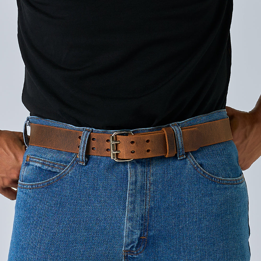 A person in a black shirt and blue jeans wears Hanks Belts’ The Legend Double Prong Retro Style leather belt with a gold double-prong roller buckle, resting their hands on their hips.