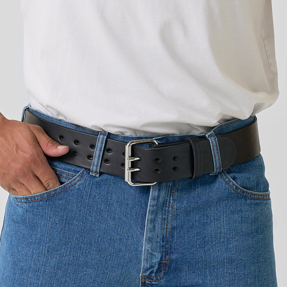 A person wearing blue jeans, a white t-shirt tucked in, and Hanks Belts' "The Marshall" black 2" wide gun belt with a silver buckle. One hand rests near the belt buckle.
