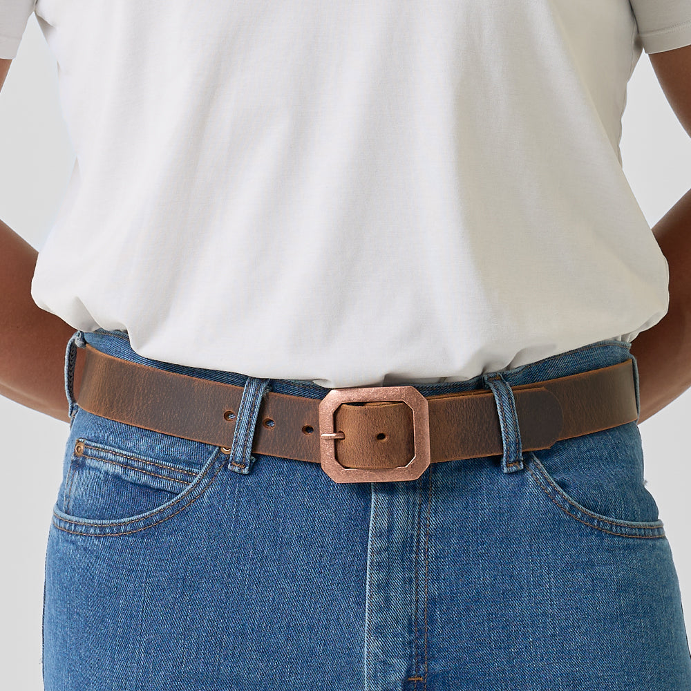 A person wears a white t-shirt tucked into blue jeans with The Relic Retro Style by Hanks Belts—a brown leather belt featuring a large, square, bronze-colored buckle.