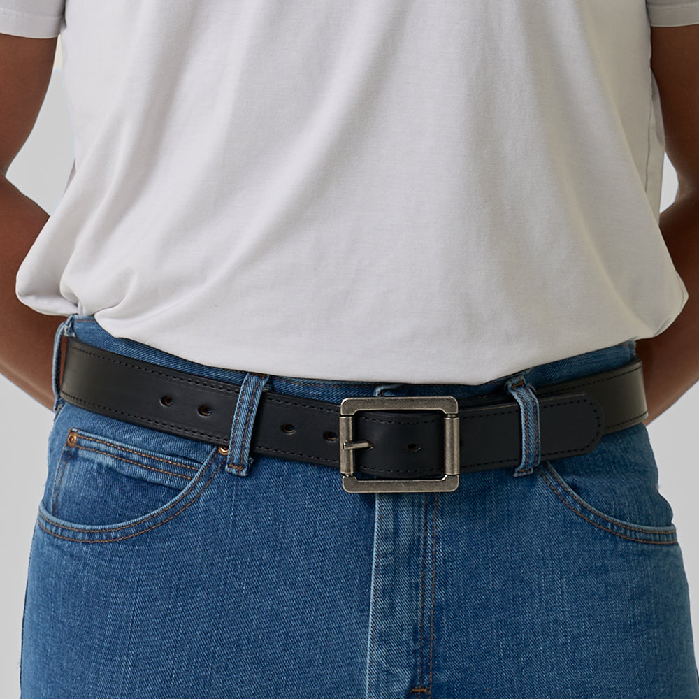 A person wearing a white t-shirt tucked into blue jeans with Hanks Belts’ The Reversible, a black leather belt with a large square silver buckle. The cropped image shows only the torso and waist.