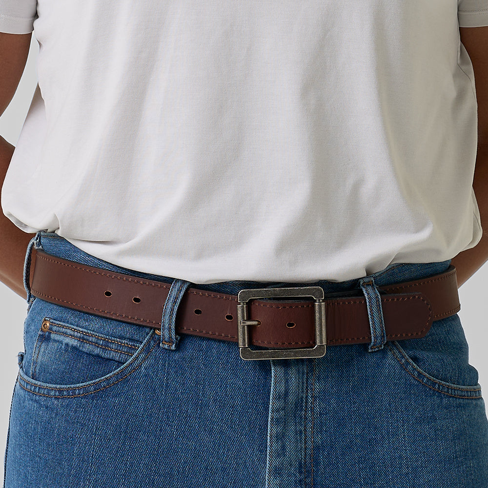 A person wearing blue jeans, a white t-shirt, and The Reversible by Hanks Belts—a reversible leather belt with a square brass buckle—shown from the waist down with hands at their sides.