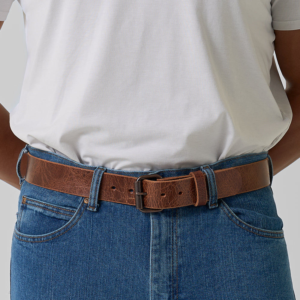 A person is seen from behind wearing a white t-shirt tucked into blue jeans, accessorized with Hanks Belts' The Rustic Glazed belt. Their hands are resting behind their back.