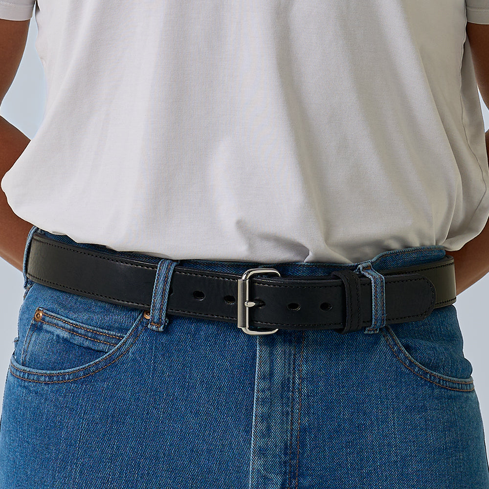 A person wears a white t-shirt tucked into blue jeans with the Hanks Belts "The Steel Core - 1.5" black leather belt, featuring a silver buckle. Their hands rest on their hips.