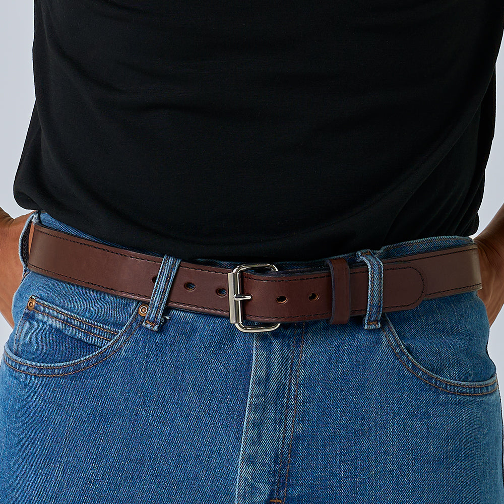 A person wears blue jeans, a black shirt, and a Hanks Belts "The Steel Core - 1.5"" brown leather gun belt with a silver buckle. The image focuses from the waist down, highlighting the CCW belt and pants.