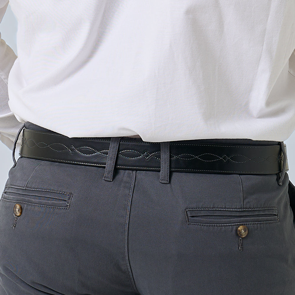 A person in a white shirt and dark gray pants wears the Hanks Belts Teton Tapered—a handcrafted leather belt with decorative stitching. The photo focuses on the waist and lower back, showcasing the artisan quality of this premium accessory.