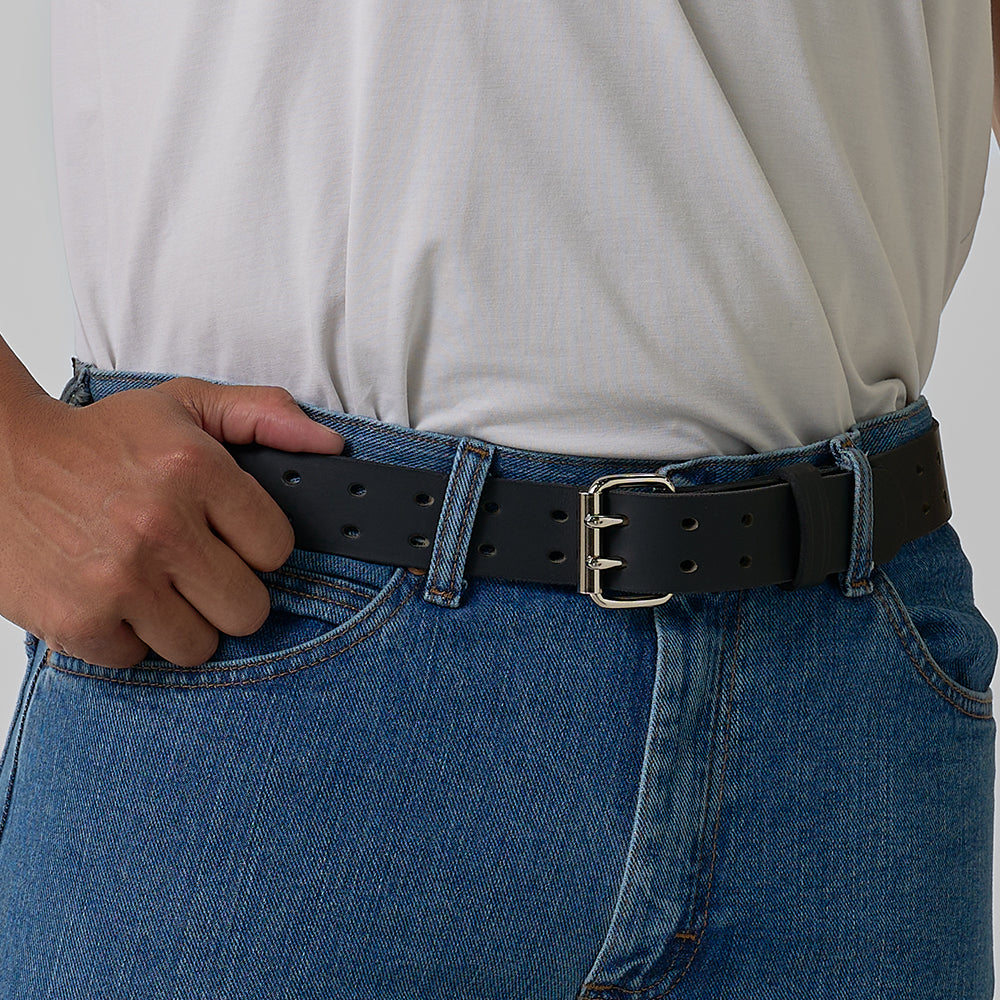 A person in blue jeans and a white shirt adjusts the Hanks Belts Woodstock Double Prong Retro Style belt with a silver buckle, highlighting the waist and vintage leather design.