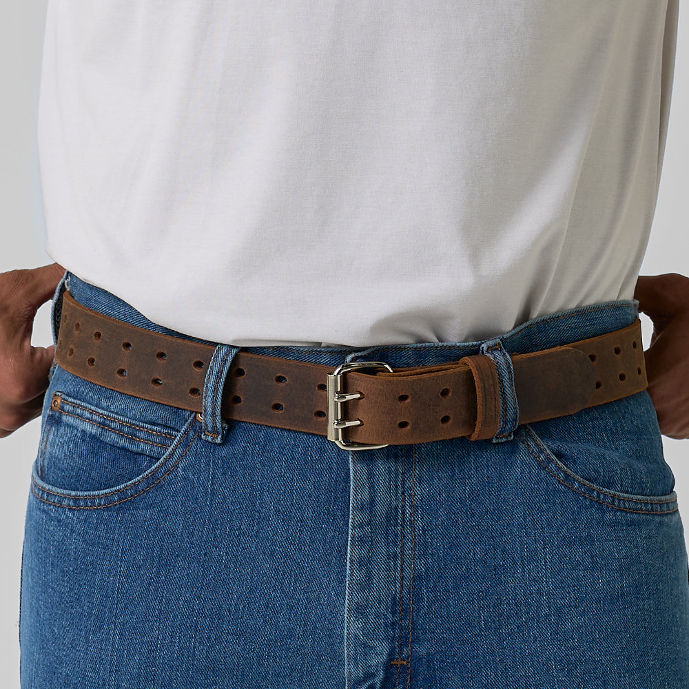 A person in blue jeans and a white tucked-in shirt wears Hanks Belts' The Woodstock Double Prong Retro Style, a full-grain leather belt with multiple holes and a silver buckle.