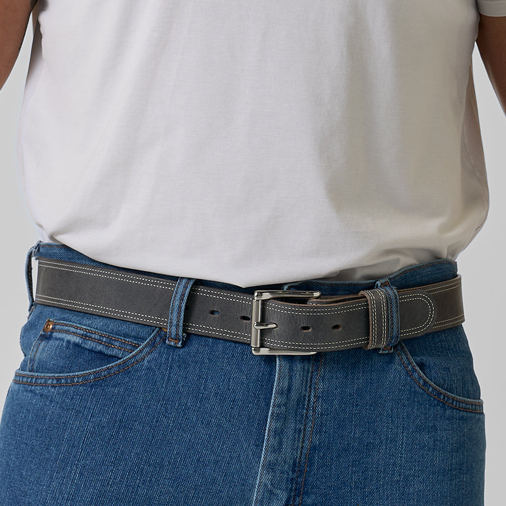 A person wears a white T-shirt, blue jeans, and The Wyoming Bison belt by Hanks Belts—crafted from bison leather with white stitching and a silver buckle. The focus is on the waist and the belt.