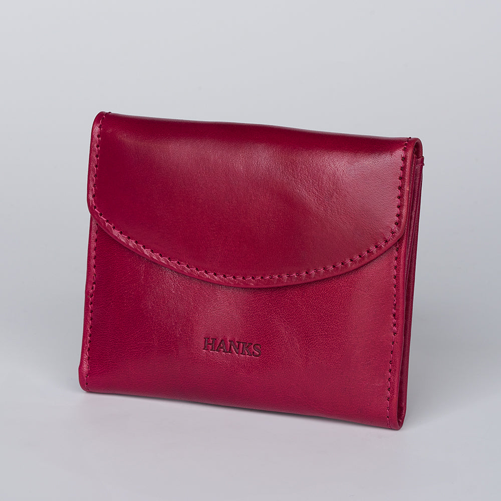 The Adelina Mini Bifold by Hanks Belts is a red leather wallet with visible stitching and "HANKS" embossed on the front, shown standing upright against a plain light gray background.