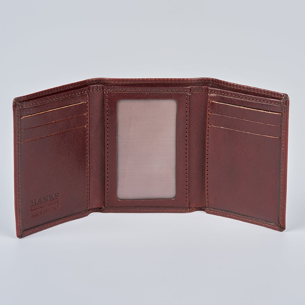 The Garda Italian Trifold with ID Window by Hanks Belts is open, revealing card slots, an ID window, and embossed "HANKS" text in the bottom left corner. It is photographed on a plain light gray background.