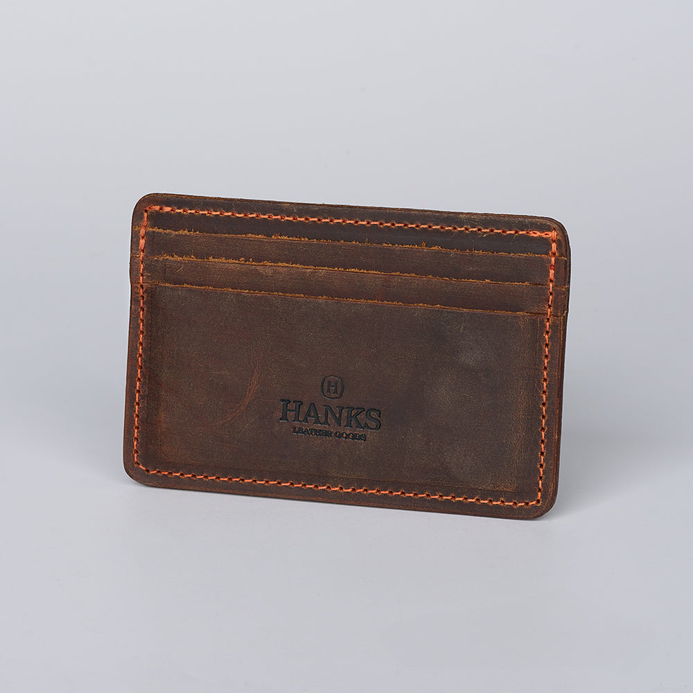 The Hamlin Card Holder with ID Window by Hanks Belts is crafted from brown steerhide leather with orange stitching, offers three card slots, a black-stamped logo on the front, and is proudly made in the USA.
