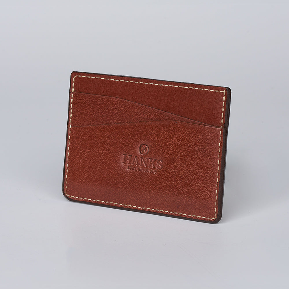 The Monroe Goatskin Card Holder by Hanks Belts features brown goatskin, white stitching, multiple card slots, and an embossed logo on the front—ideal as a sleek, minimalist wallet. Displayed against a plain light background.