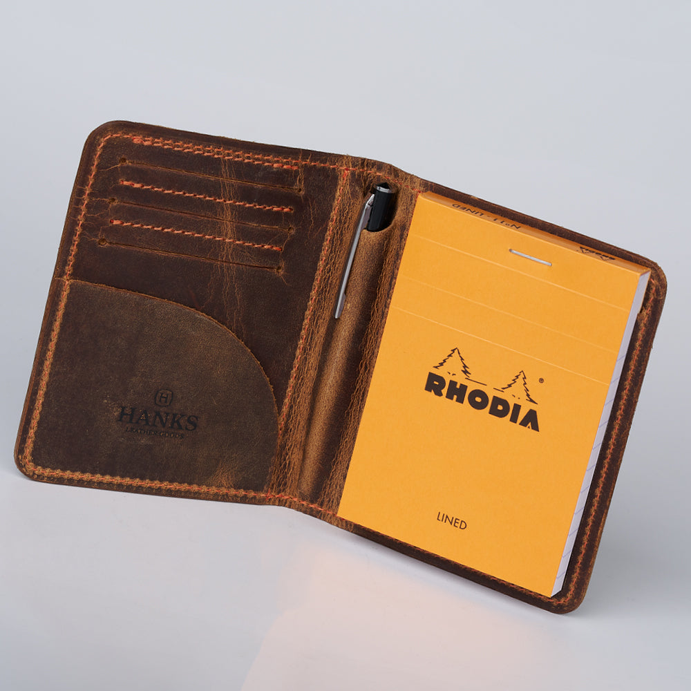 The Hanks Belts Notesman and Credit Card Wallet is a brown, handcrafted leather wallet featuring a yellow Rhodia notepad and pen loop on the right, with card slots and a pocket on the left.