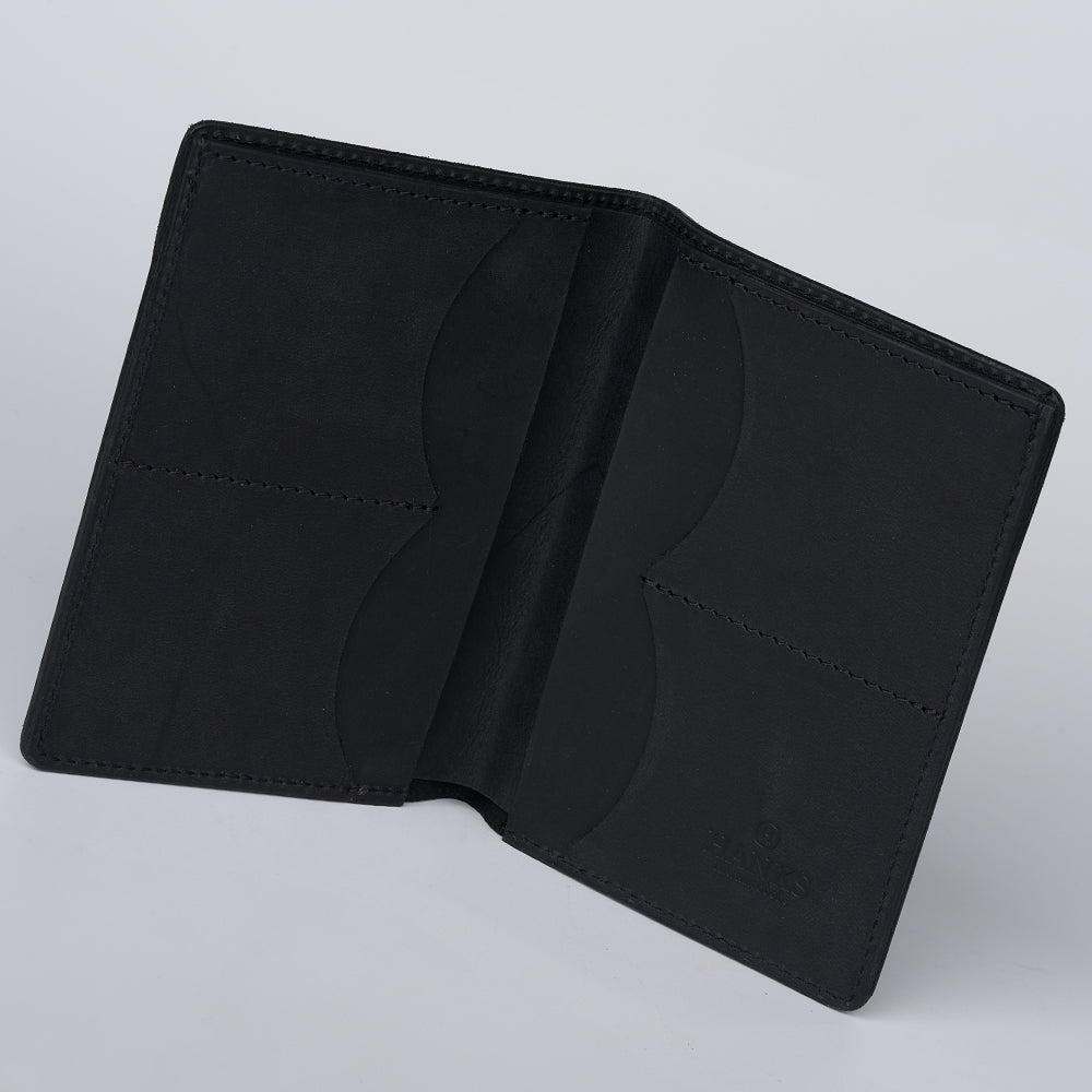 A Hanks Belts Passport Case Wallet, crafted from North American Steerhide in black leather with multiple card slots and clean edge stitching, is displayed empty on a light gray surface.