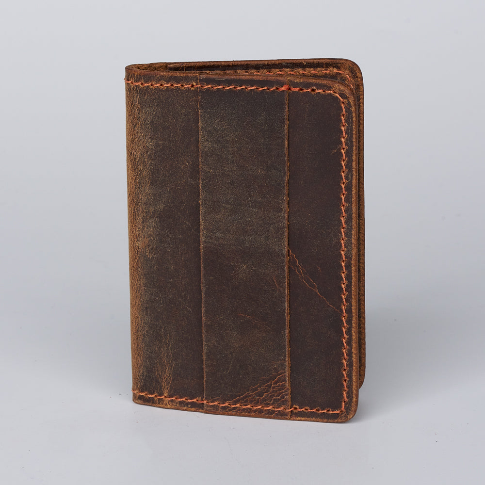 The Hanks Belts Tupper Bifold Card Holder, a brown USA-made wallet with visible stitching and a distressed leather finish, stands upright closed on a simple light surface.