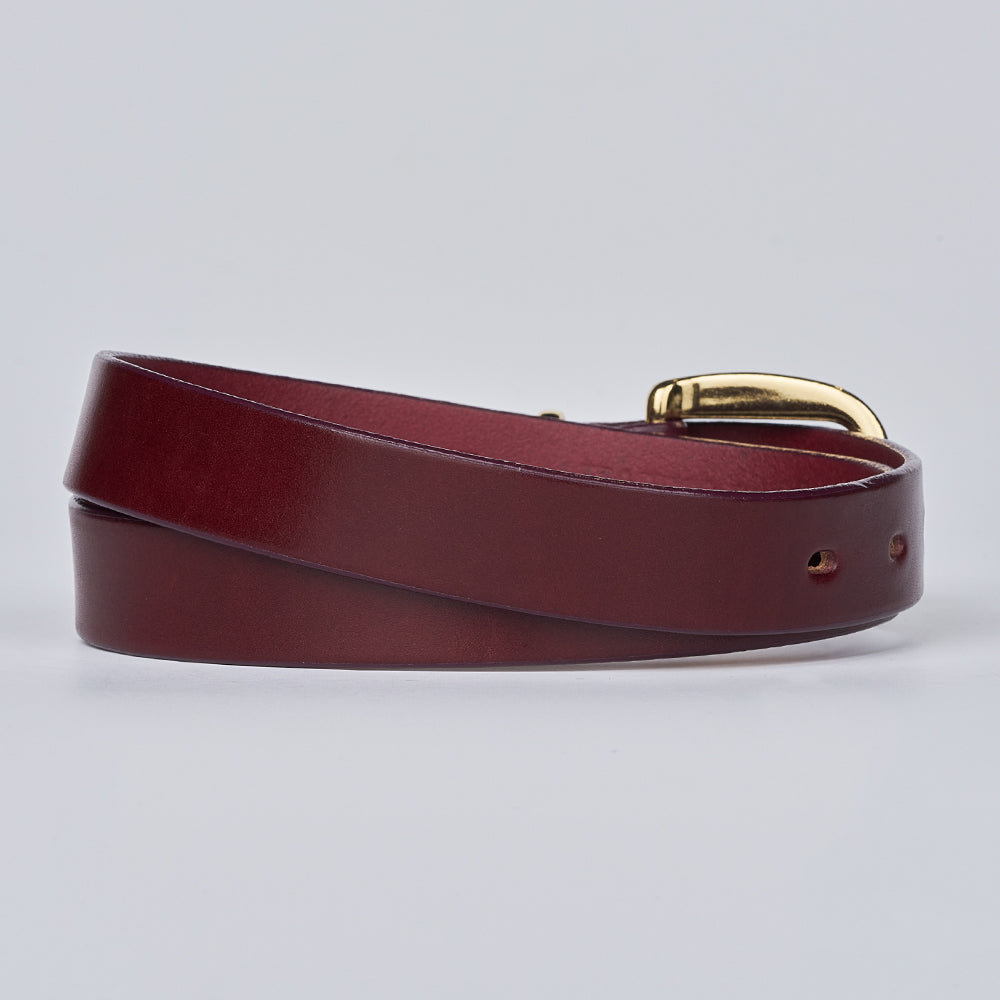 The Poppy by Hanks Belts is a rolled-up burgundy women's leather belt with a shiny brass buckle, shown on a light gray background—ideal for adding a statement touch to any outfit.