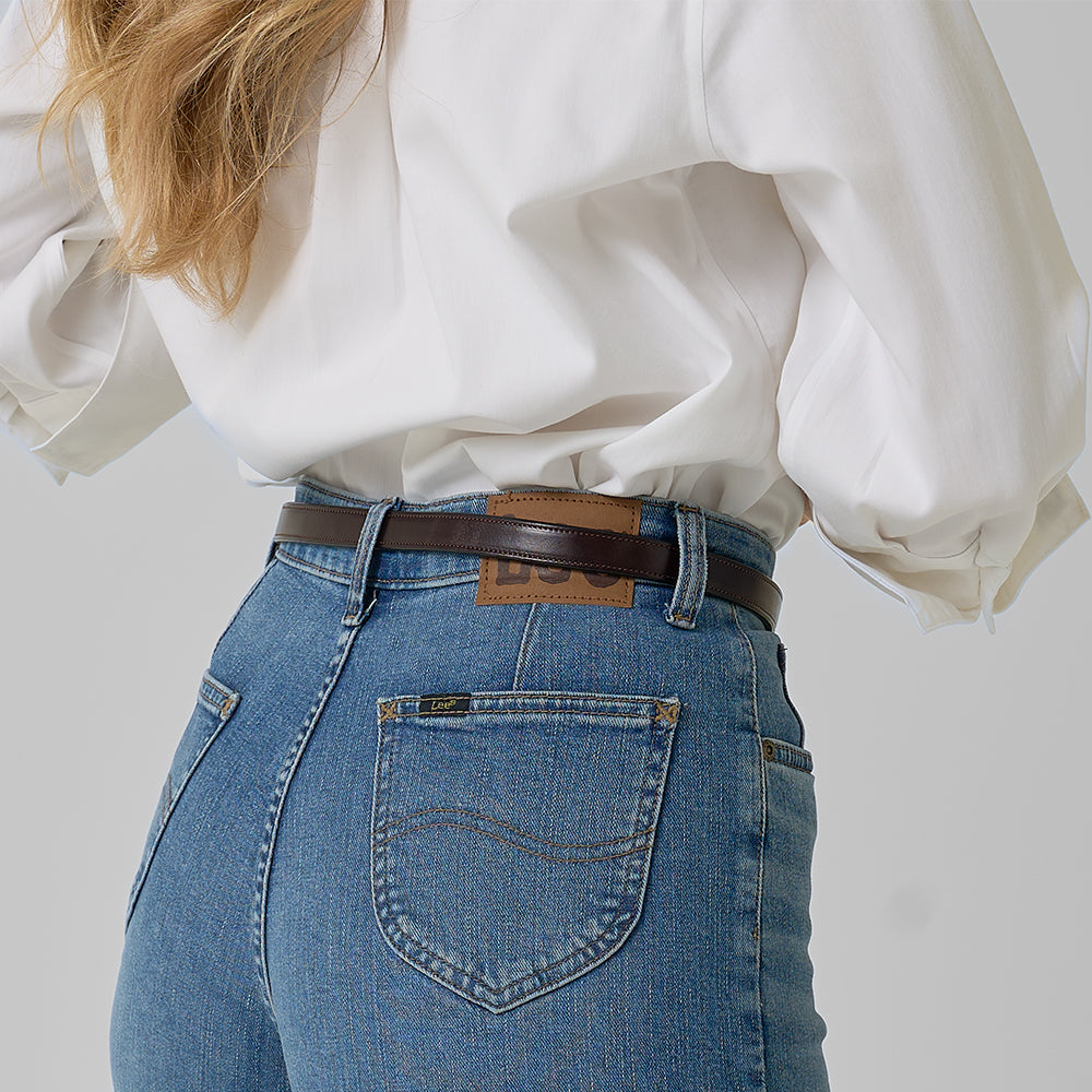 A person with long, wavy blonde hair wears a white blouse and high-waisted blue jeans, styled with Hanks Belts' The Dixie women's leather belt crafted from English Bridle leather. The photo is taken from behind against a plain background.