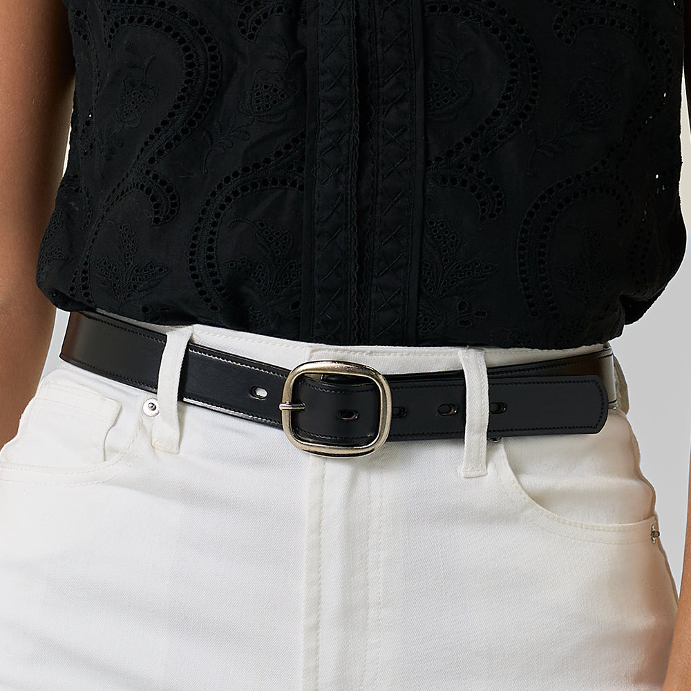 Wearing a black textured blouse with high-waisted white pants, they accessorize with The Eloise women's leather belt by Hanks Belts, made in the USA and finished with a silver buckle. Their hands rest partially in their pockets.