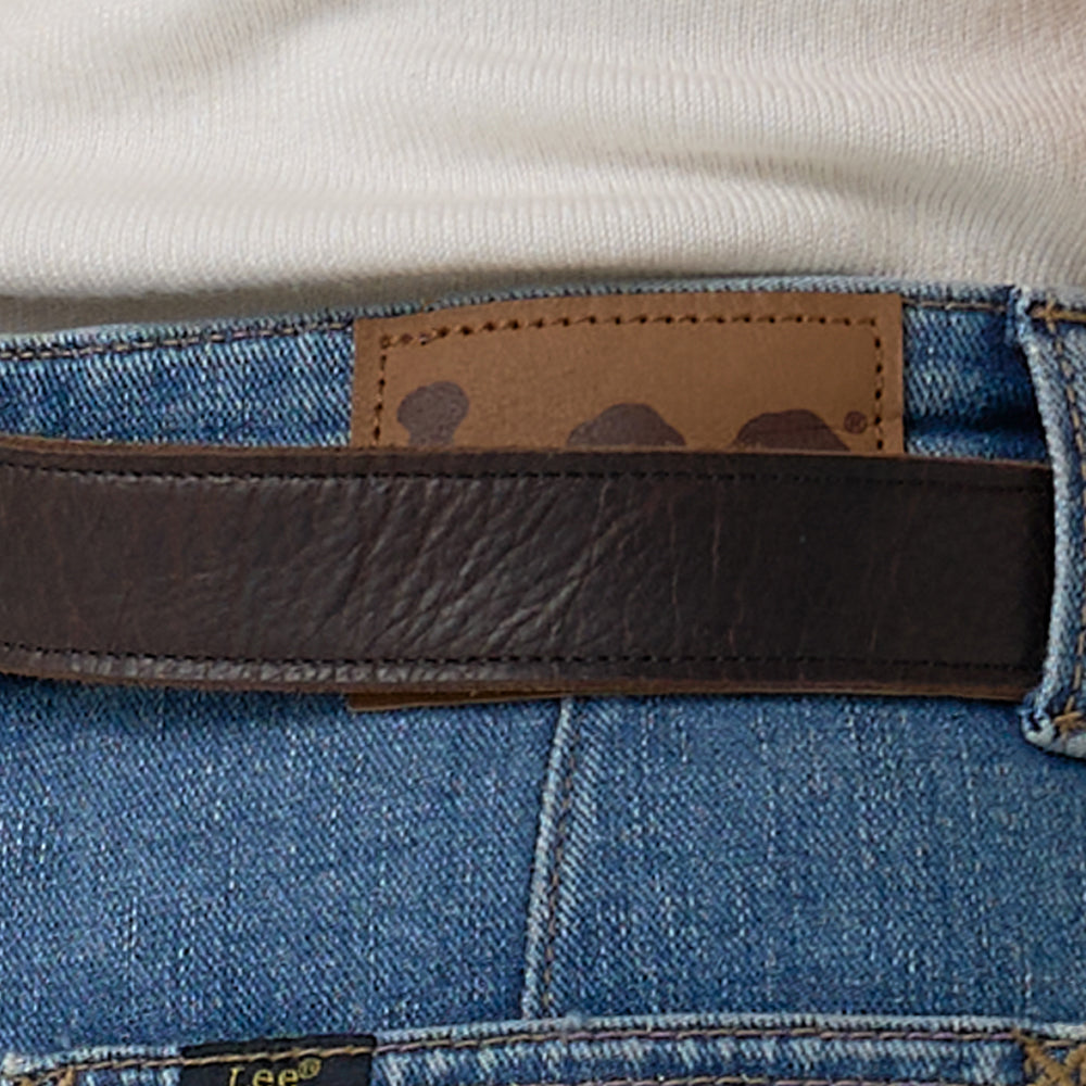 Close-up of a person wearing Hanks Belts' Ember - 1.25" brown leather belt with blue jeans and a white shirt, showing part of a rectangular brown patch on the jeans' waistband.