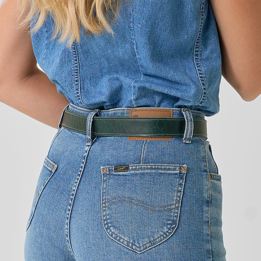 A person in blue jeans and a sleeveless denim shirt, seen from behind, wears the Hanks Belts Ember - 1.25" black belt with a brown patch on the waistband. Their blonde hair hangs over one shoulder.